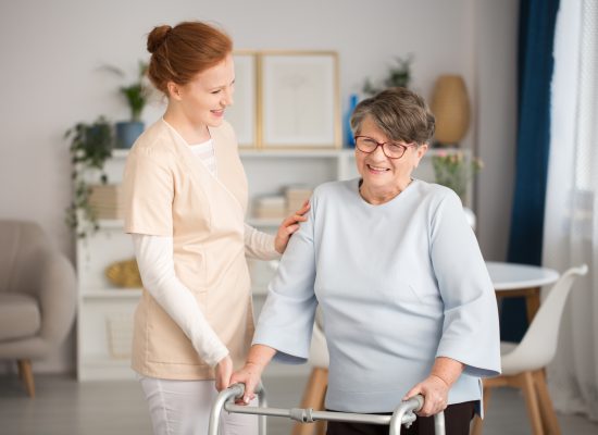 Professional medical caretaker in uniform helping smiling senior woman with a walker in a living room of private luxury healthcare clinic